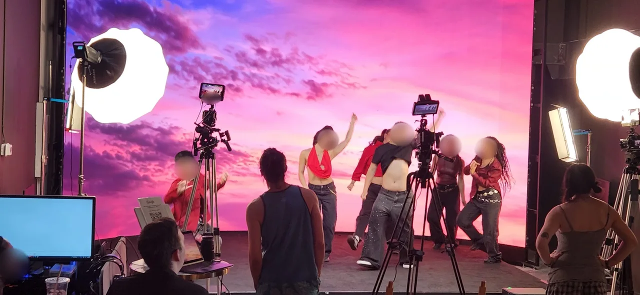 Dancers in red tops mid-jump during a dance video shoot against a pink sky backdrop