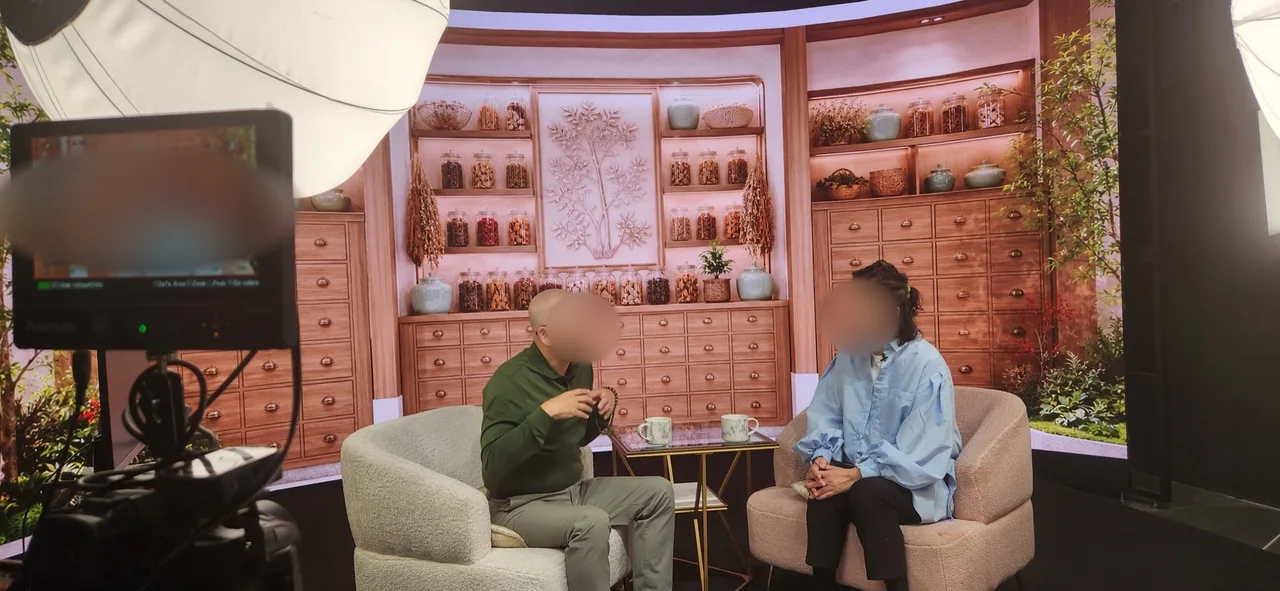 Talk show host in a green shirt interviewing a guest in blue in front of a traditional Chinese pharmacy LED backdrop