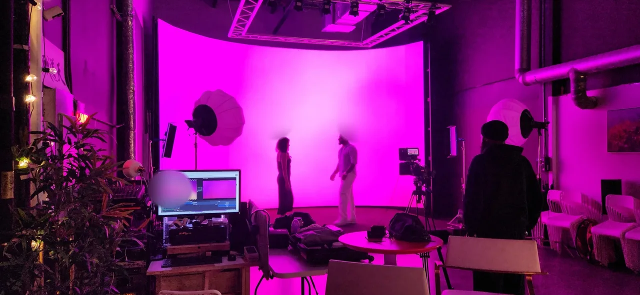 Wide behind-the-scenes shot of a music video set bathed in magenta light with singer and co-star