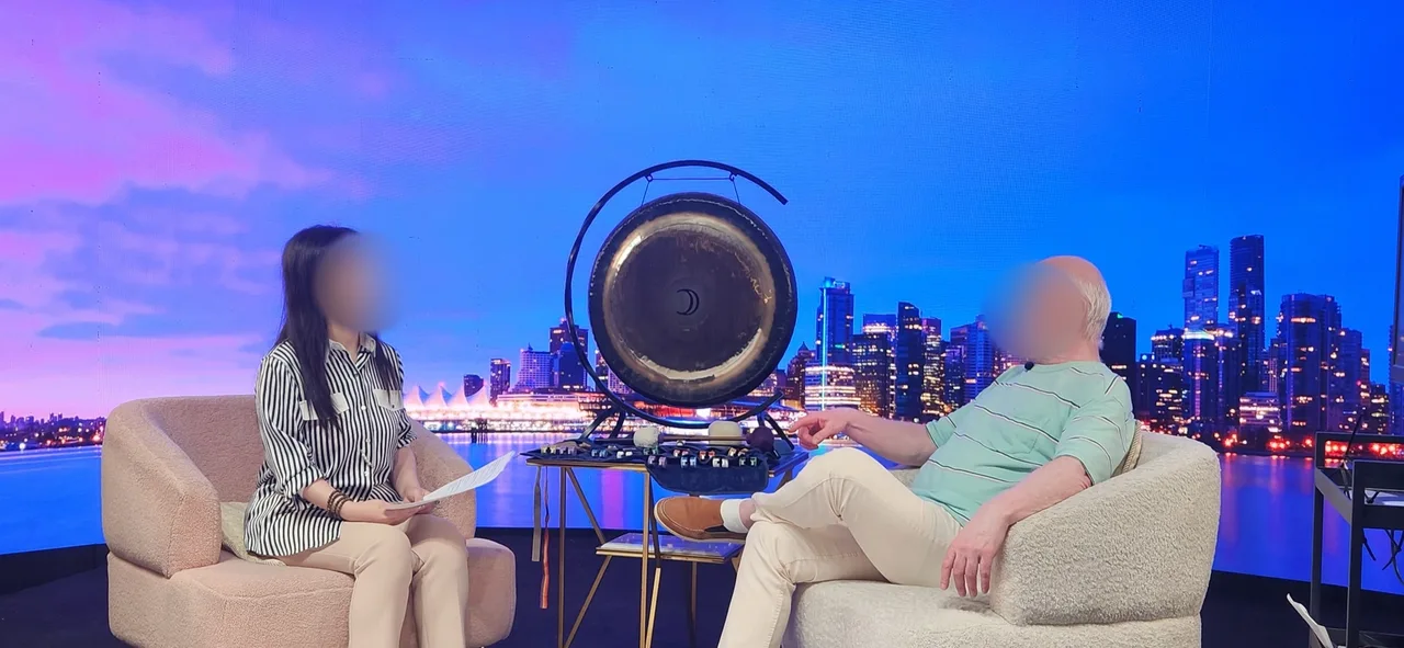 Host with papers interviewing a bearded guest beside a large gong with the Vancouver skyline on the LED wall