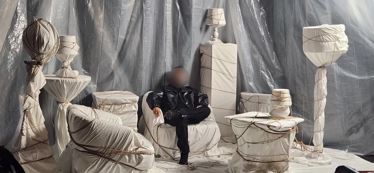 Singer in a black leather jacket seated among rope-bound white furniture on an art-directed set