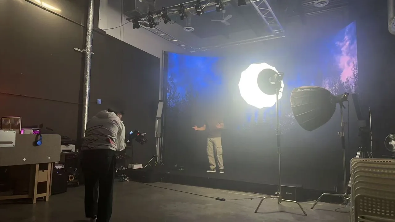 Singer performing with open arms in front of an LED wall showing a blue sky and pine forest, cinematographer filming with a gimbal in the foreground
