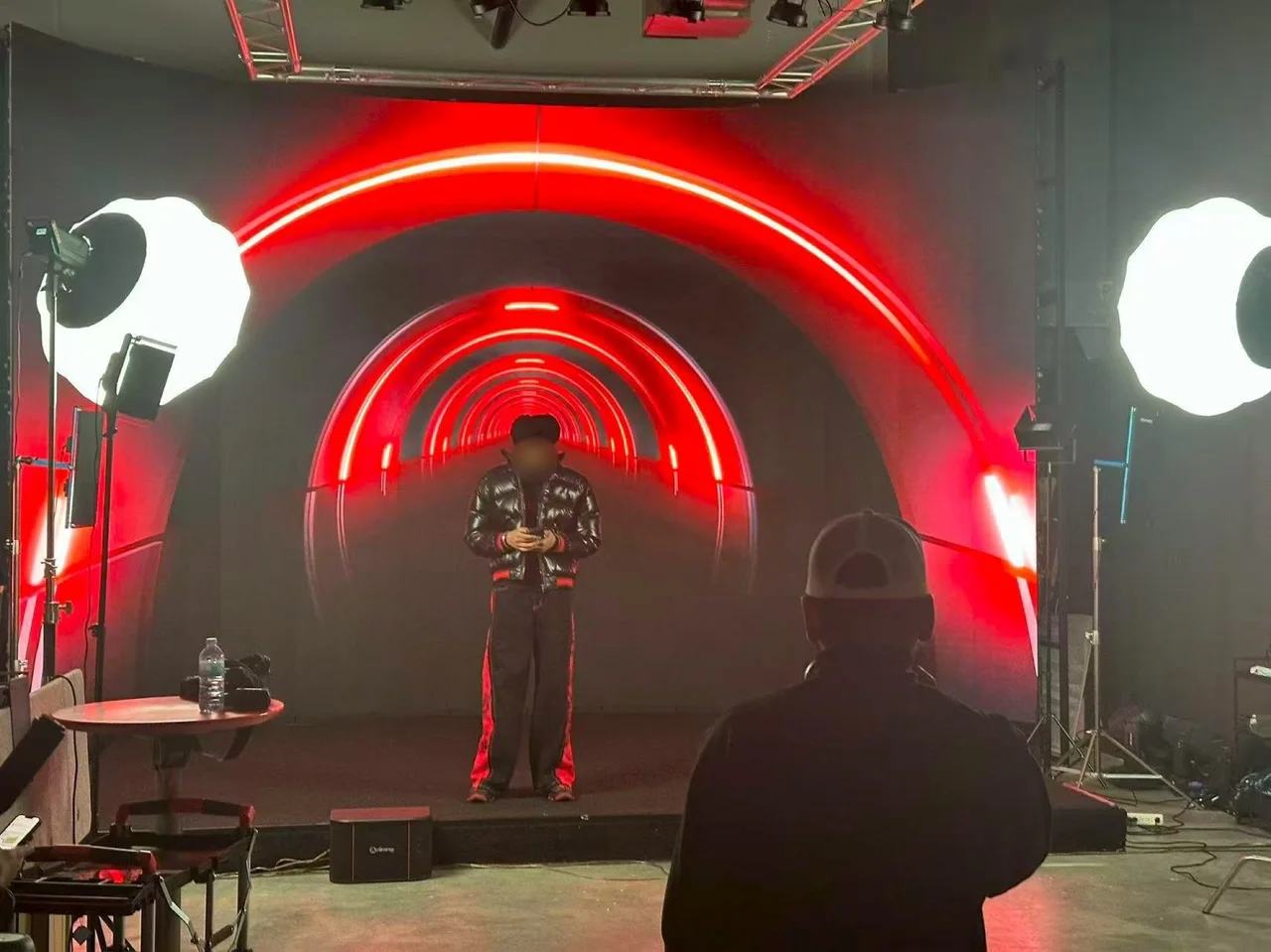 Punjabi singer in a black turban and glossy puffer jacket standing in a red neon tunnel displayed on the LED wall at Upperland Studio, director watching from the foreground