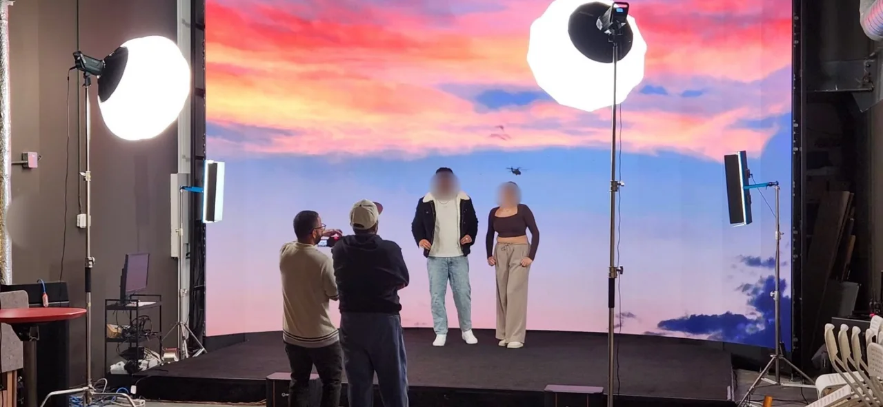 Lead actor and actress standing before an LED wall sunset sky with a helicopter silhouette during the Route 66 Films music video shoot, filmed by a camera operator