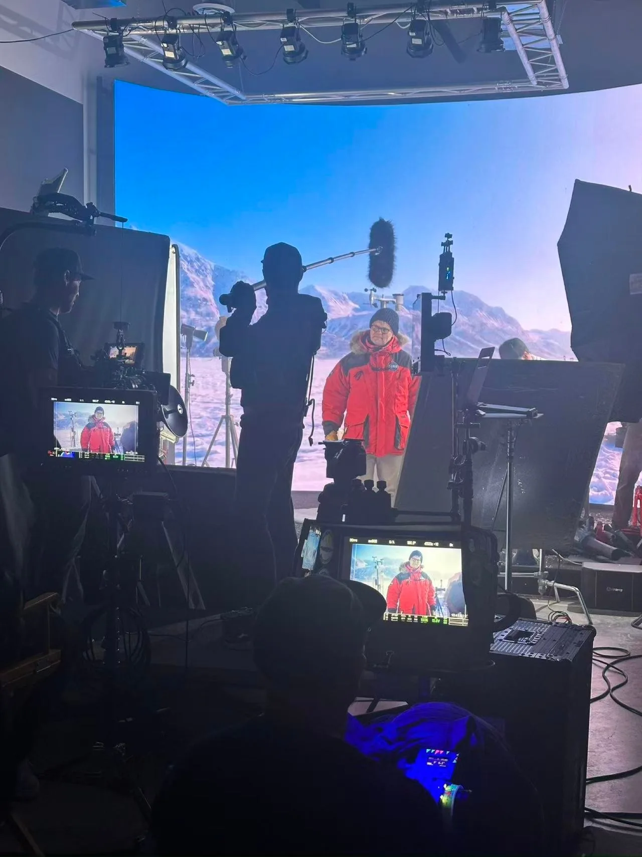 A presenter in a red AERIS jacket before the Arctic LED mountain backdrop, with boom microphone and camera monitors at Upperland Studio